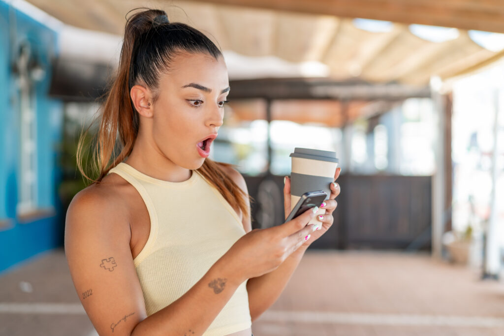 El Poder del Café 1 young pretty woman outdoors using mobile phone holding coffee with surprised expression